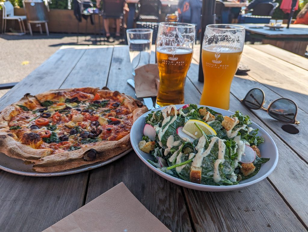 A plate of salad next to a pizza and two glasses of beer on a wooden picnic  table.