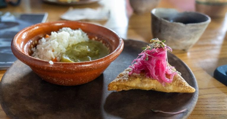A close up photo of a black plate on a table with a small pastry next to a brown bowl with rice and a green sauce inside it.