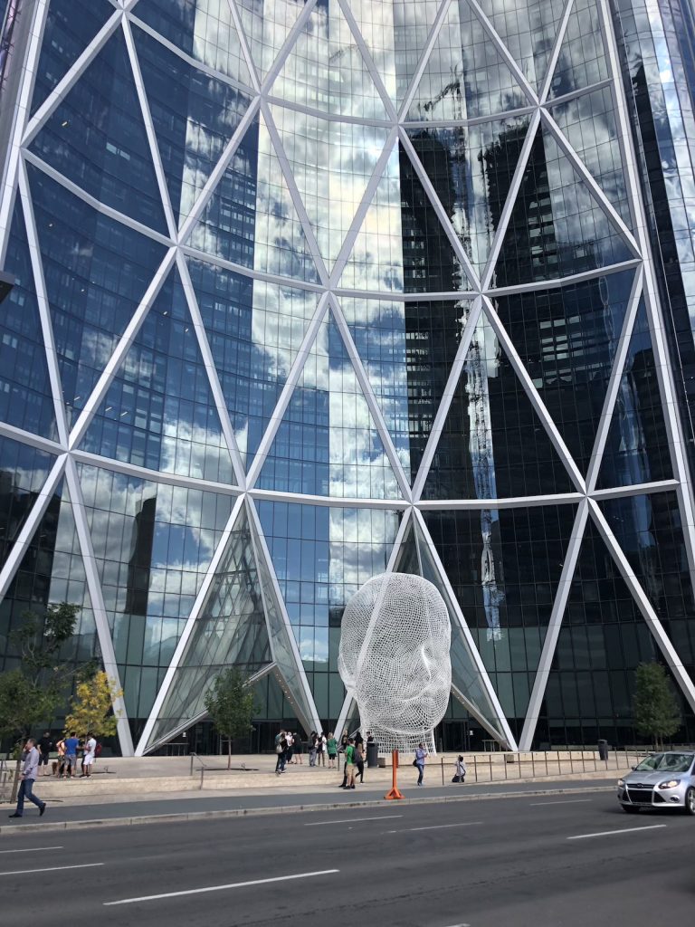 A large, white head sculpture made from wire, towered over by a glass building.