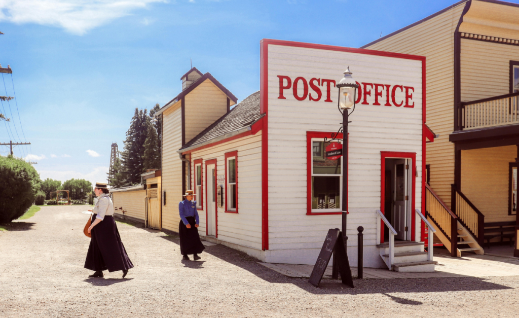 Two women in old-fashioned costumes outside a wooden historic post office on a sunny day.
