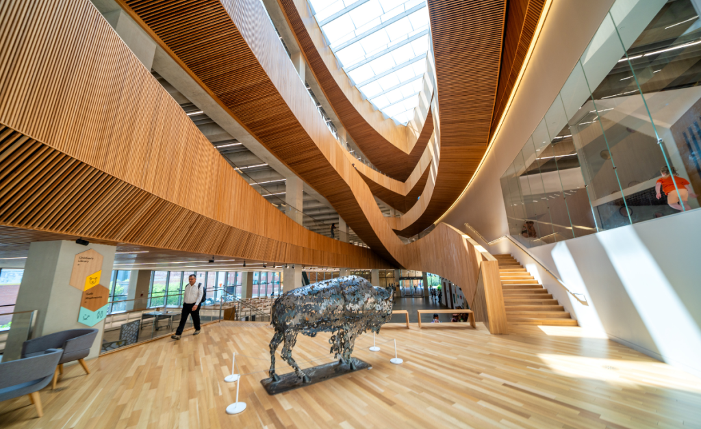 A metal bison sculpture inside a wooden building with different levels leading to a window in the ceiling.