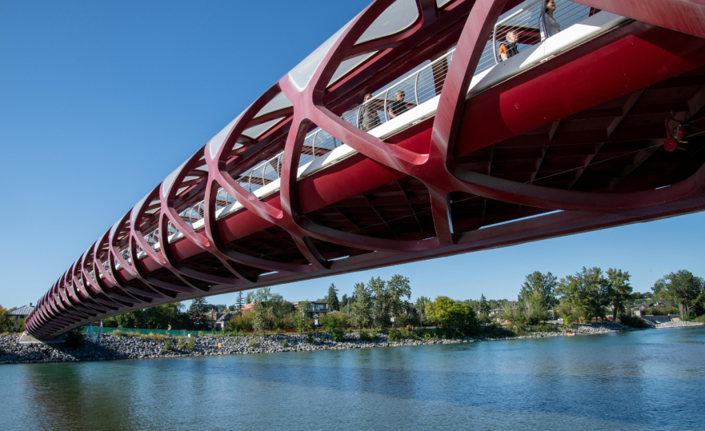 A close-up view of the red Calgary Peace Bridge taken from below the bridge, showing the river underneath and clear blue sky above.