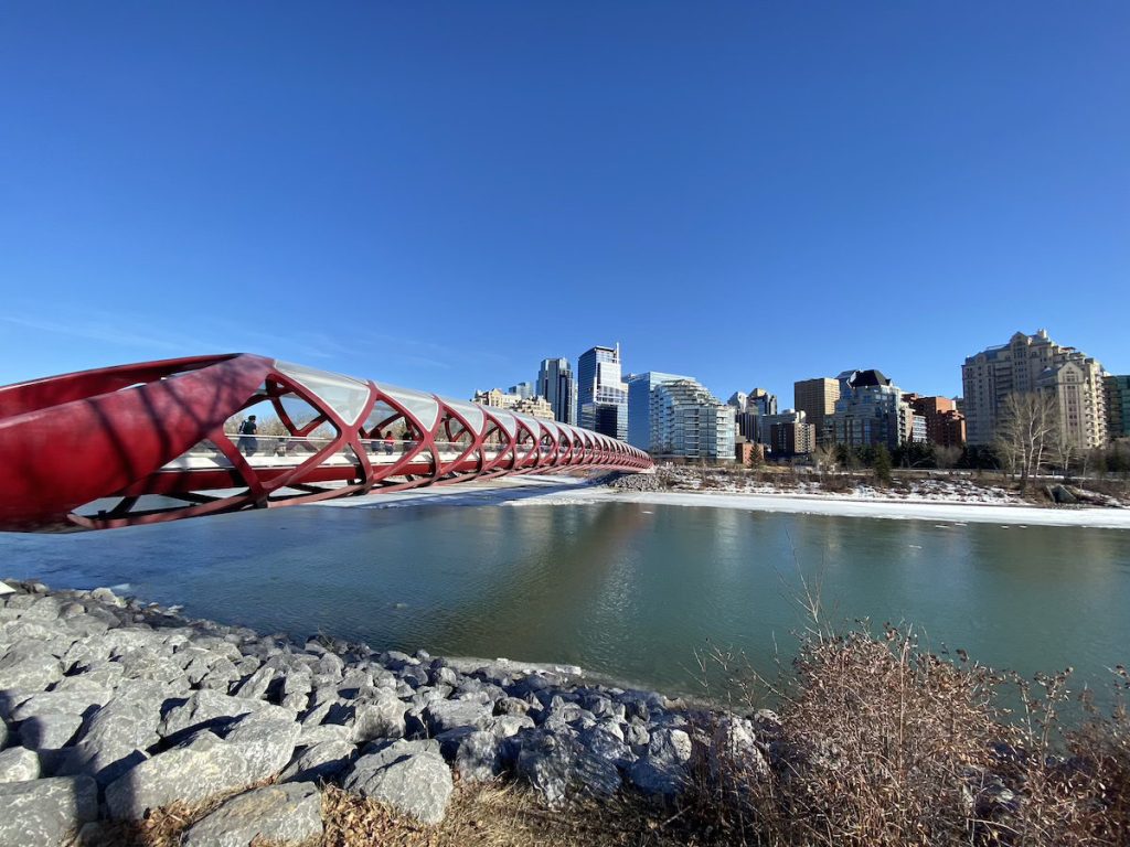 The red Calgary Peace Bridge stretching across the river with snow on the opposite bank and a clear blue sky above.
