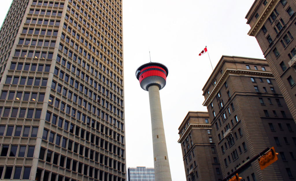 A tall tower lit up with red next to just as tall and taller buildings, one of which is flying a Canadian flag.