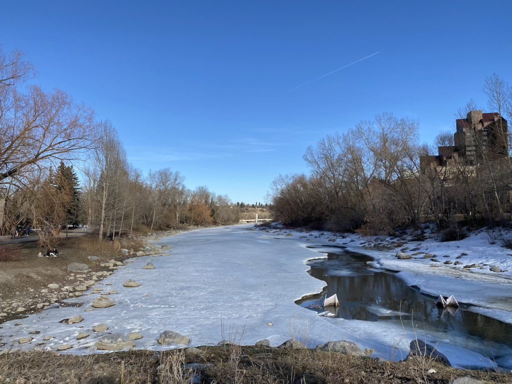 A wide river almost completely frozen over under a clear blue sky with bare trees on either side.