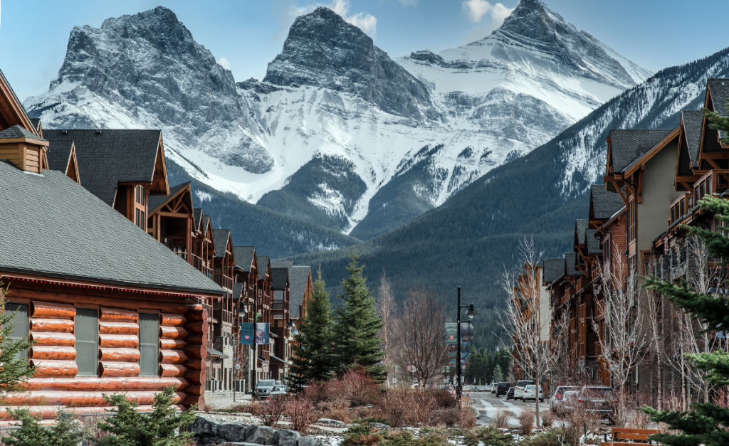 Wooden buildings and pine trees line a slightly snowy street with three huge mountain peaks covered in snow in the background that look very close.