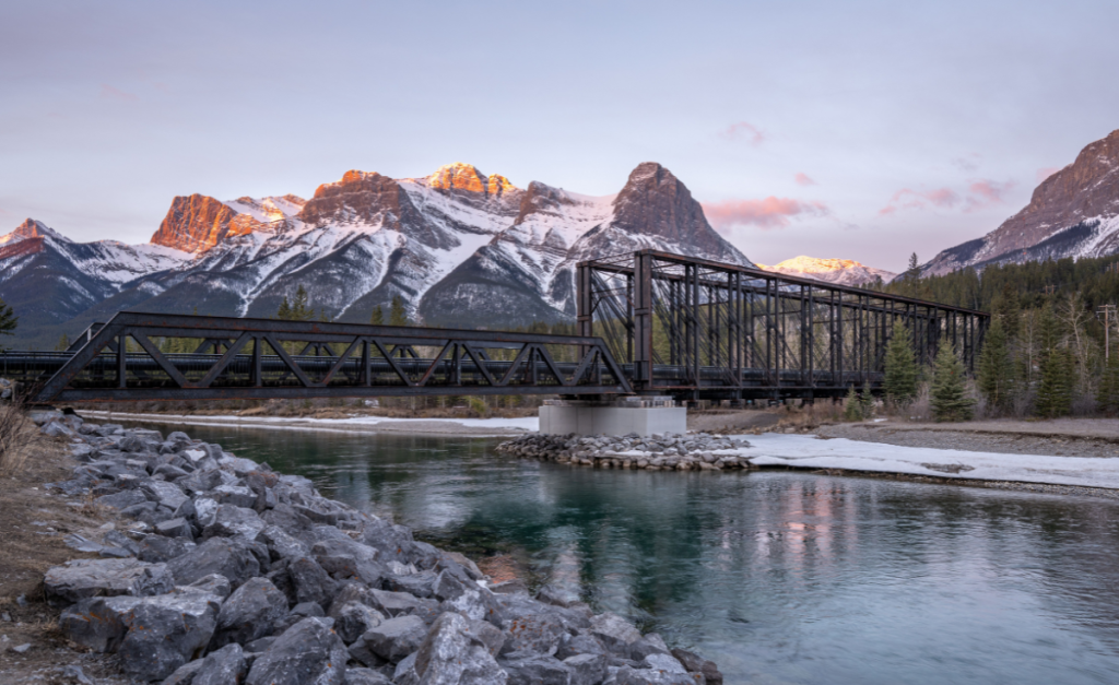 A two-level iron bridge across a rocky river, with snow-capped mountains in the background.