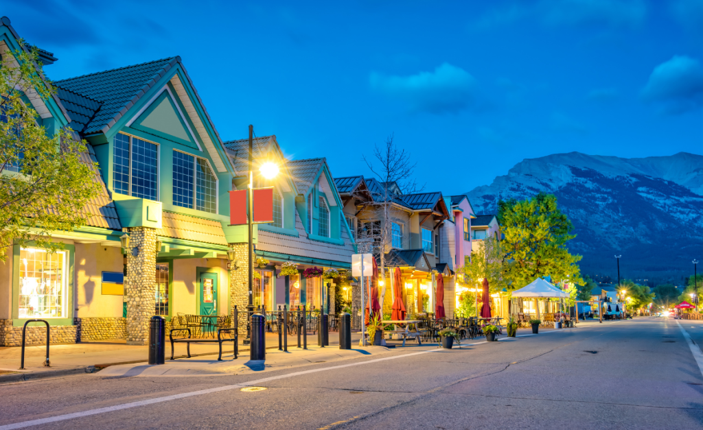 An empty street lined with shops and street lights at dusk, with mountains in the distance.