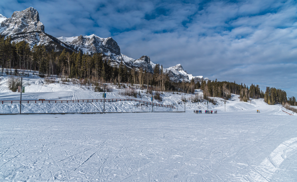 A flat open area of snow with a group of people in the distance and pine trees with snowy mountain peaks in the background.