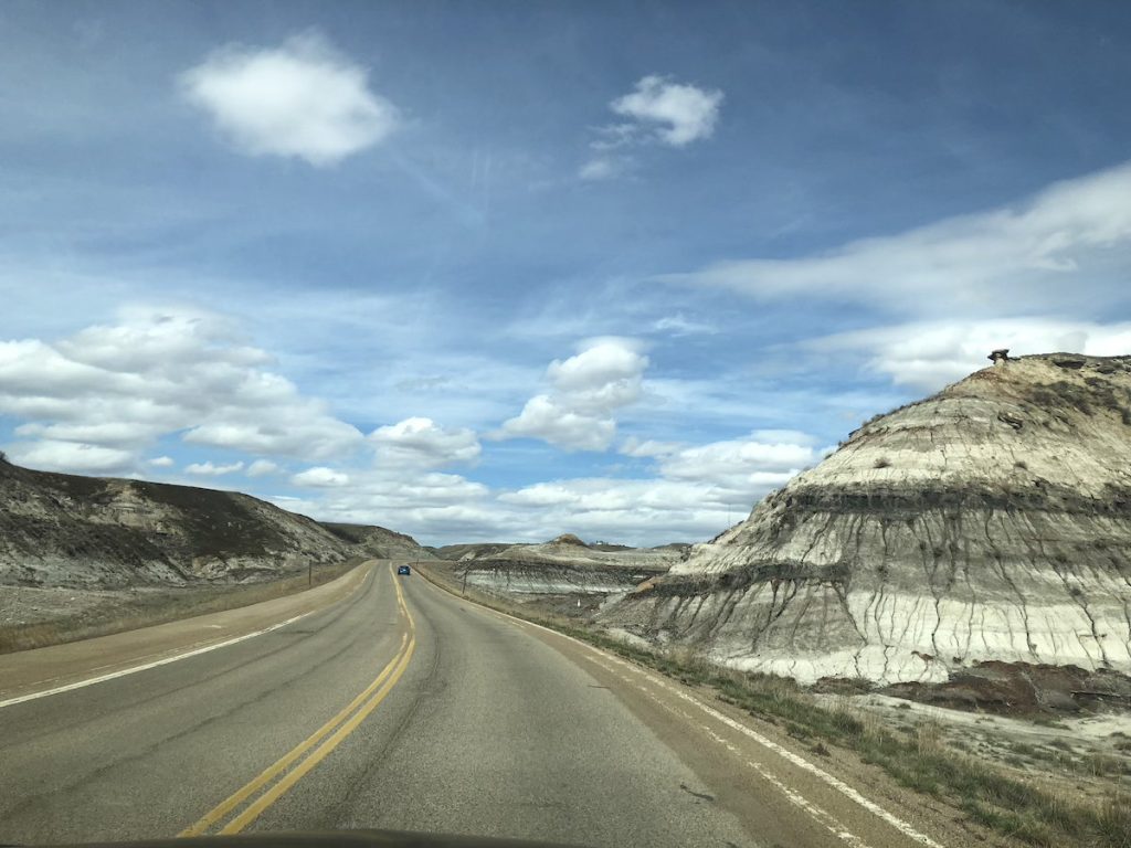 A road with one car driving among low hills under a blue sky with fluffy white clouds.