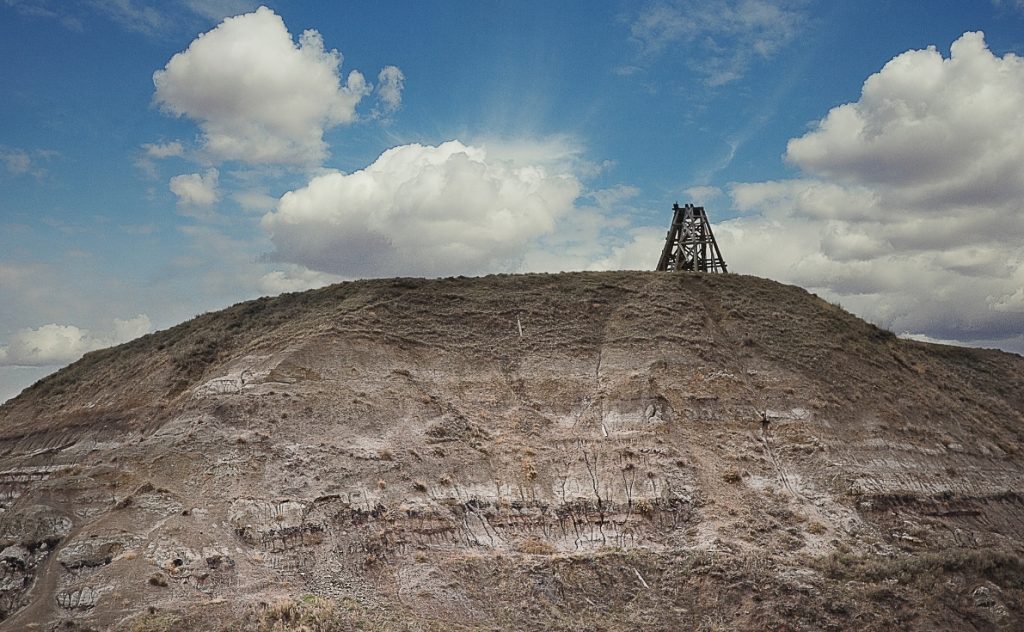 A rocky hill with a wooden structure on top and a blue sky with fluffy white clouds above.