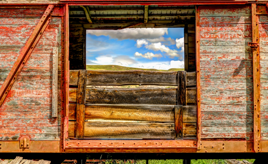 A close-up view of a weathered mine cart looking through a window to the other side where there is a hill and blue sky with fluffy white clouds.