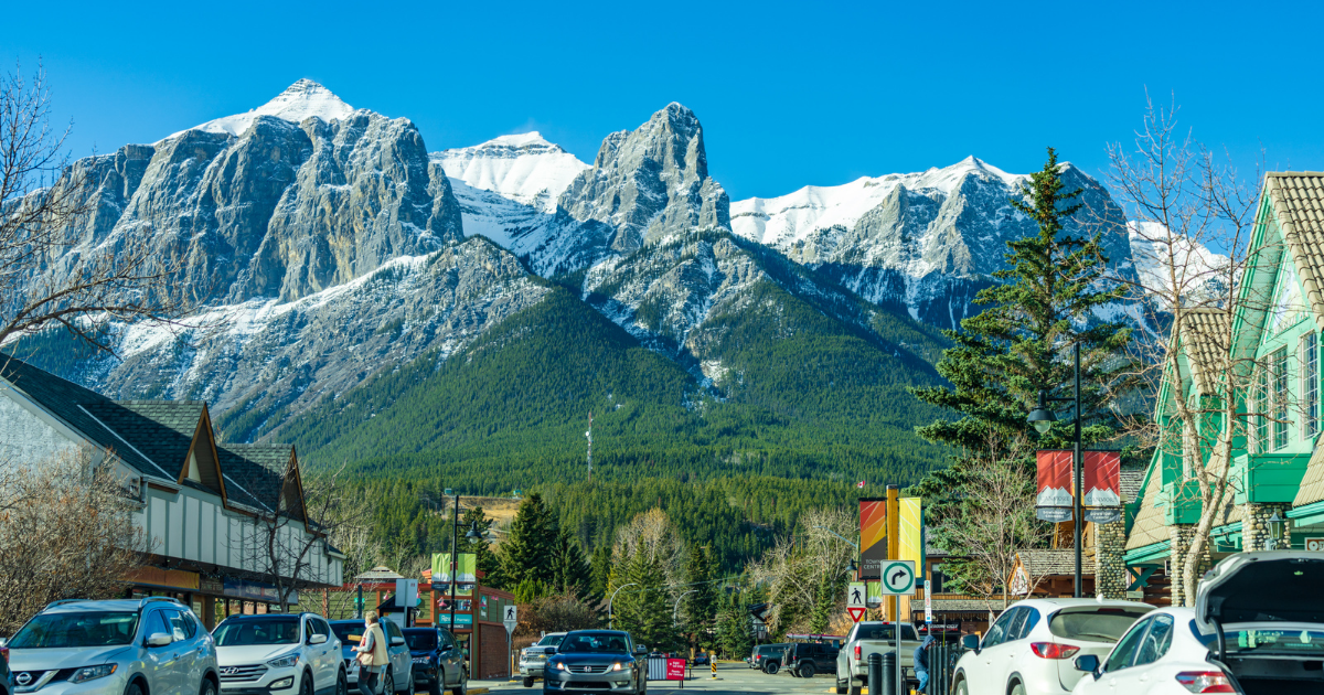 A wide street lined with cars leading to a mountain with three peaks capped in snow under a cloudless sky.