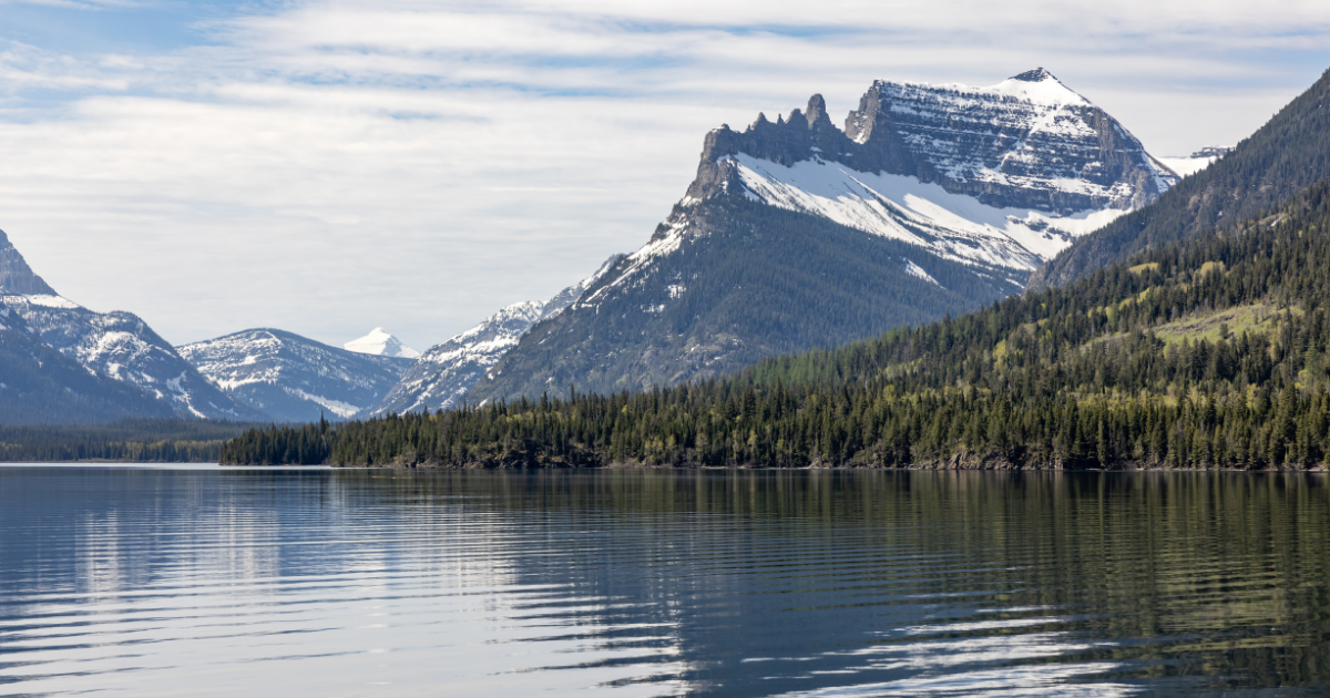 A jagged mountain with some snow on the peaks behind a hill covered in pine trees and a smooth lake in the foreground.