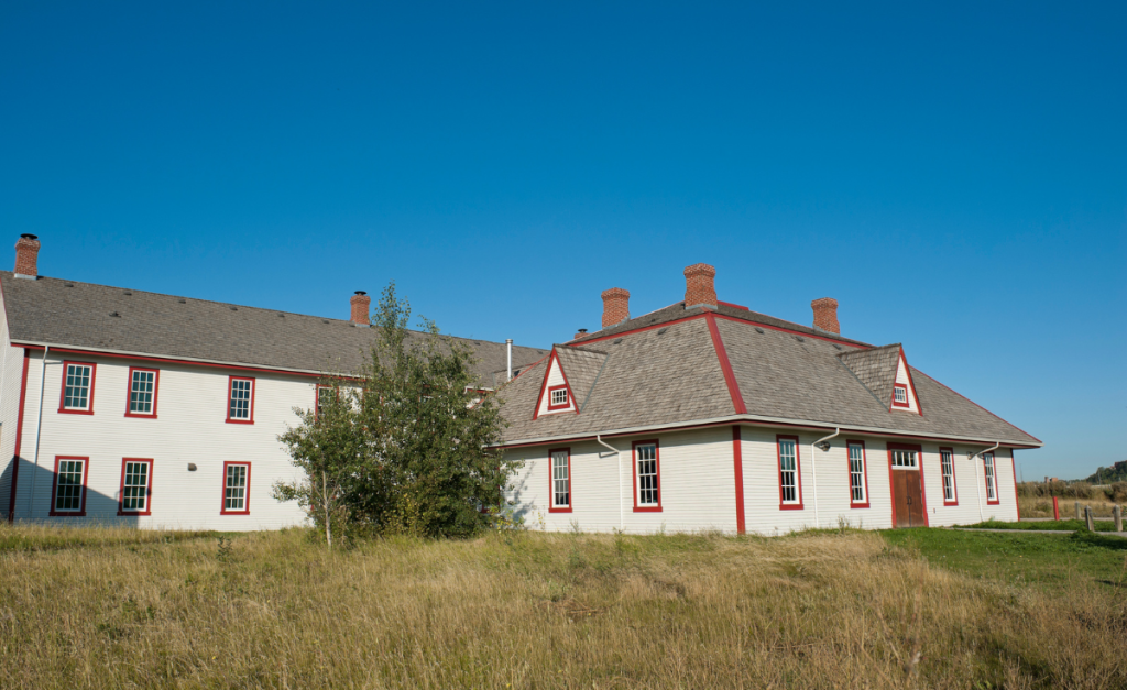Two white wooden buildings with red trim sitting on a field with a clear blue sky behind.