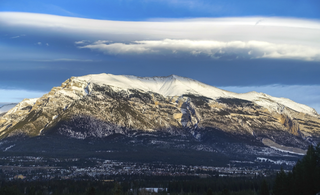 A mountain covered with snow with small buildings and pine trees at the foot and a long cloud formation above.