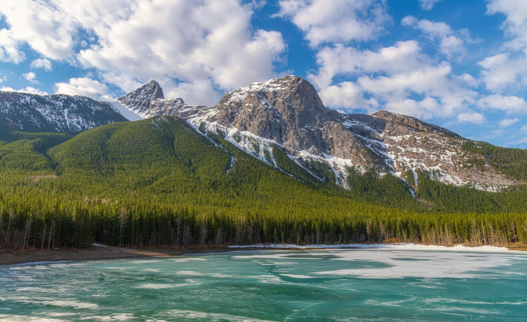 A landscape photo of a frozen aquamarine lake, with pine trees rising from the banks to rocky mountains with snow on top and blue sky with fluffy white clouds above.