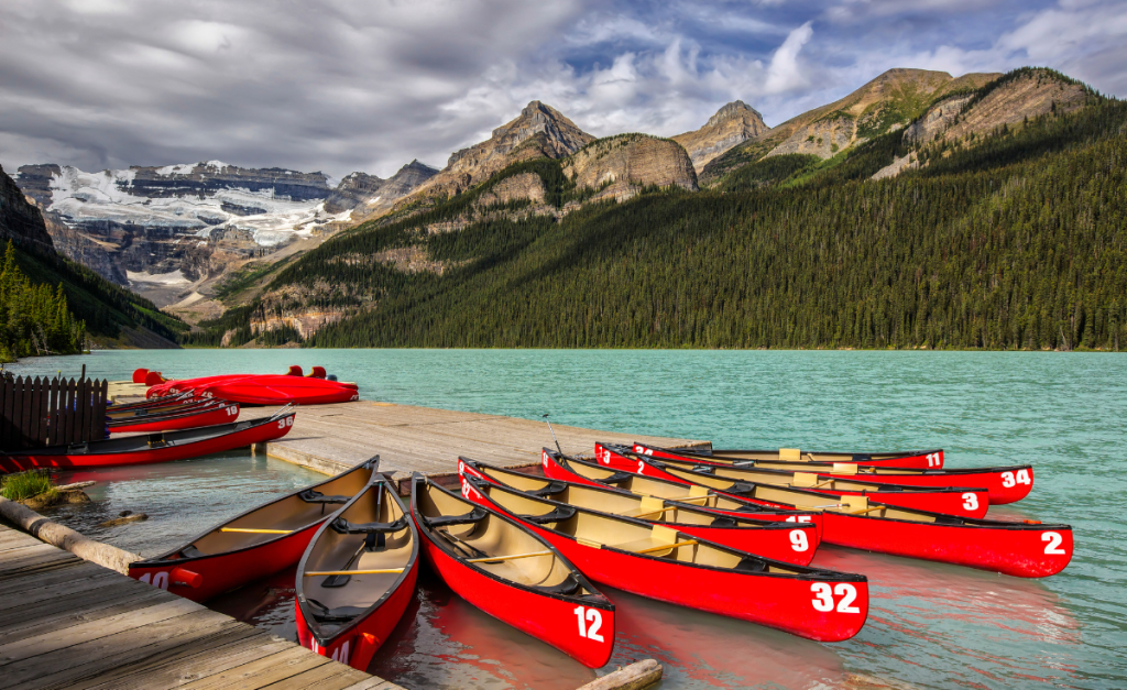 A group of red canoes docked at a wooden jetty on a light blue lake with mountains and pine trees in the background.