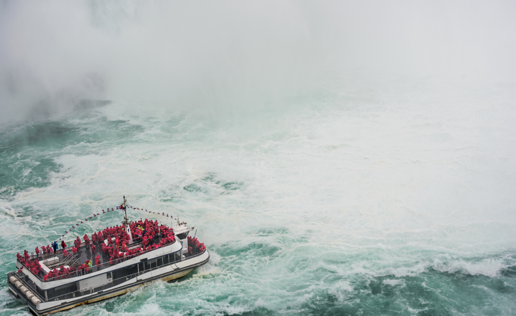 A close up shot of a cruise boat with lots of people with red raincoats on at the base of a waterfall with lots of white foam.