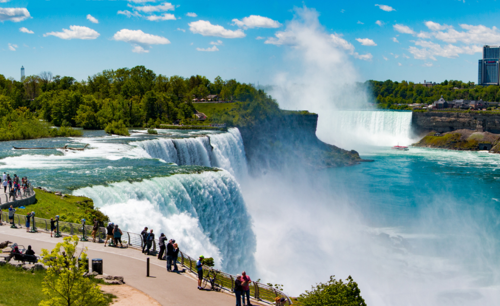 People standing on a pathway overlooking Niagara Falls from the Canadian side on a sunny day.