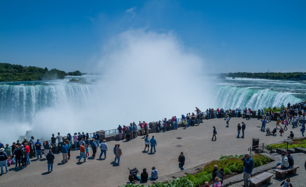Lots of people on a viewing platform overlooking the top of Horseshoe Falls as Niagara Falls, with a clear blue sky above.
