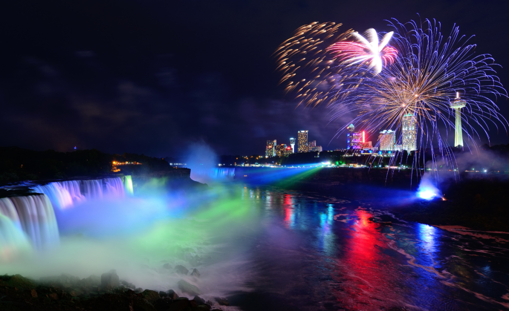 Niagara Falls at night with illuminated lights on the falls and fireworks in the sky above the buildings on the river bank.