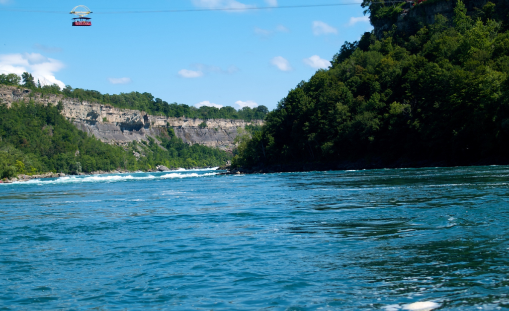 A red cable car looking tiny suspended above a wide river with rocky cliffs on one side and trees on the other.
