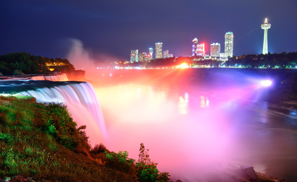 Niagara Falls at night with colored lights illuminating the falls from buildings lit up on the riverbank.