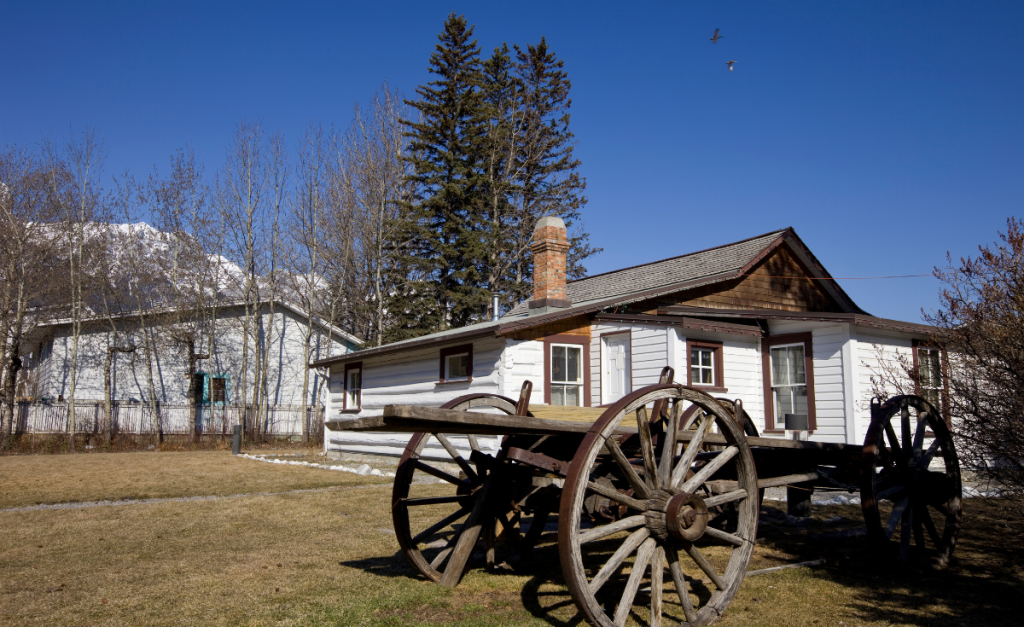 A white, wooden building with an old wooden cart in front of it and clear blue skies above.