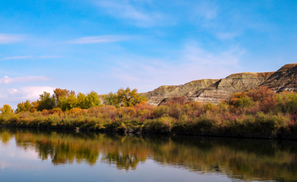 A river lined with fall colored trees and bushes with rocky hills and blue sky in the background.