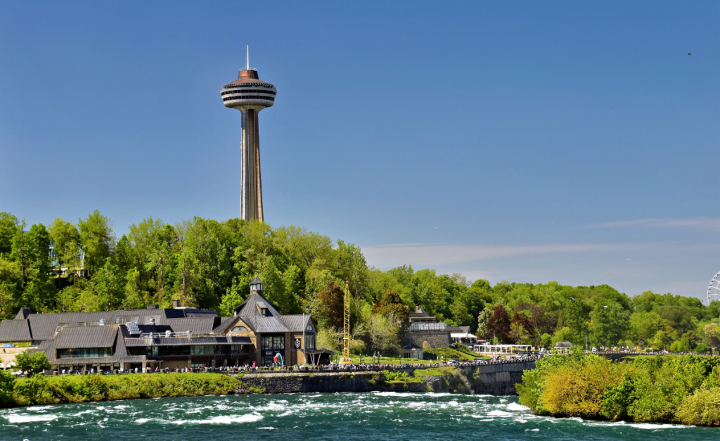 Skylon Tower at Niagara Falls rising up from behind trees and buildings on the river bank on a clear sunny day.