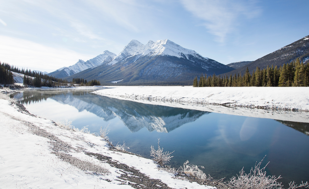 A completely still river flanked by snowy banks and reflecting the snow-capped mountain peaks in the background.