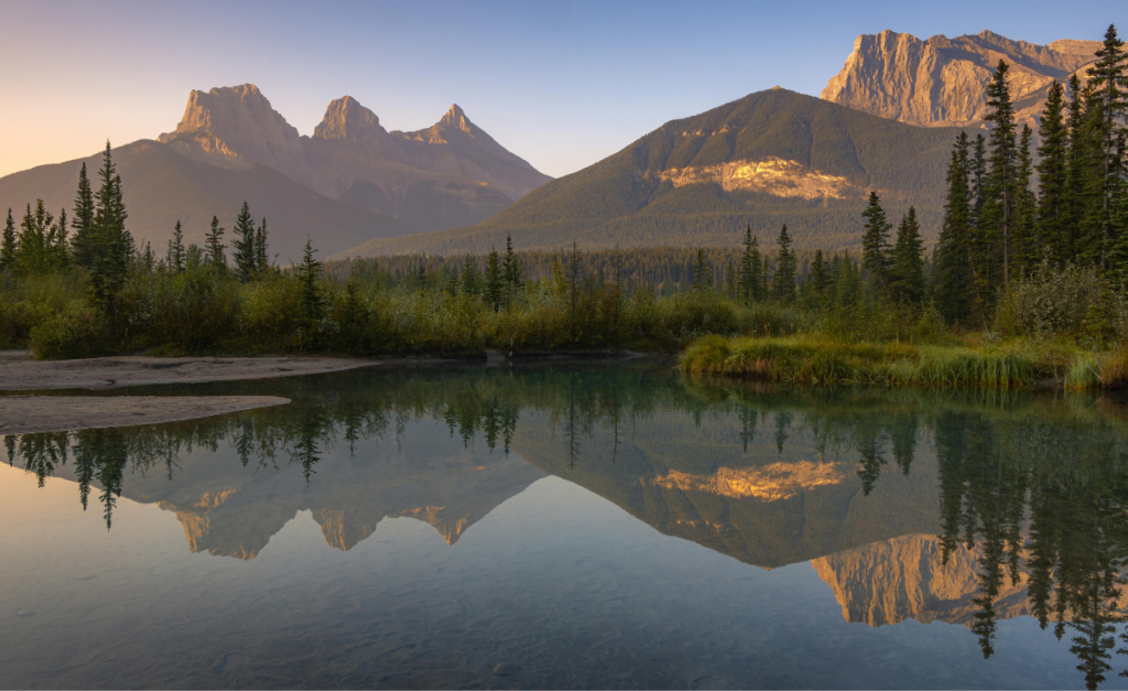 A mountain range with three distinct peaks reflecting into calm water during golden hour.