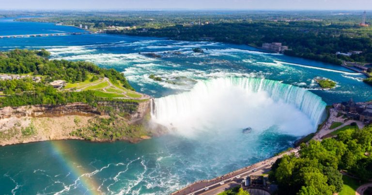 A huge curved waterfall seen from the sky with a boat at the base, buildings and trees on either side of the river and a small piece of a rainbow in the foreground.