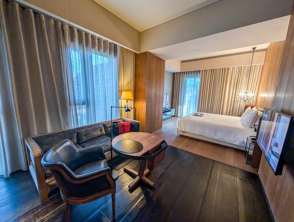 A spacious hotel suite with a couch, chair and table facing a TV, and a bed facing another TV beyond.