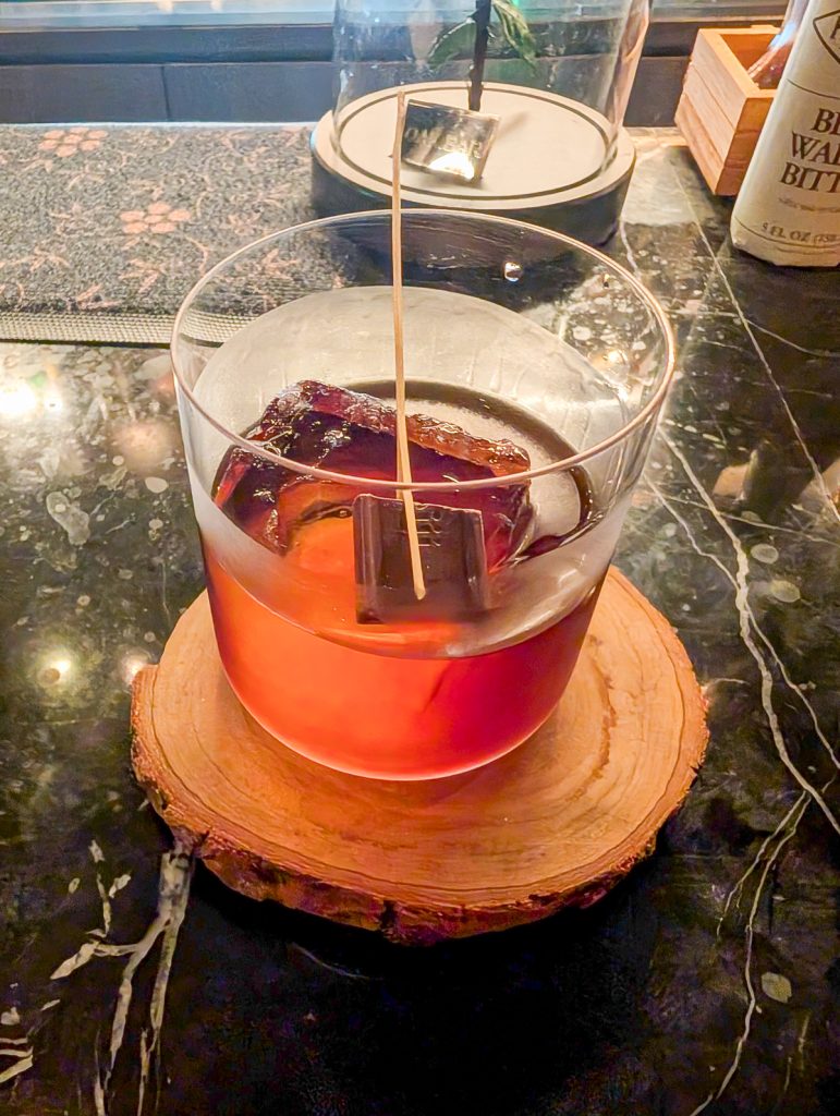 A close-up shot of a red drink with a piece of chocolate in it, sitting on a wooden coaster on a black bartop.