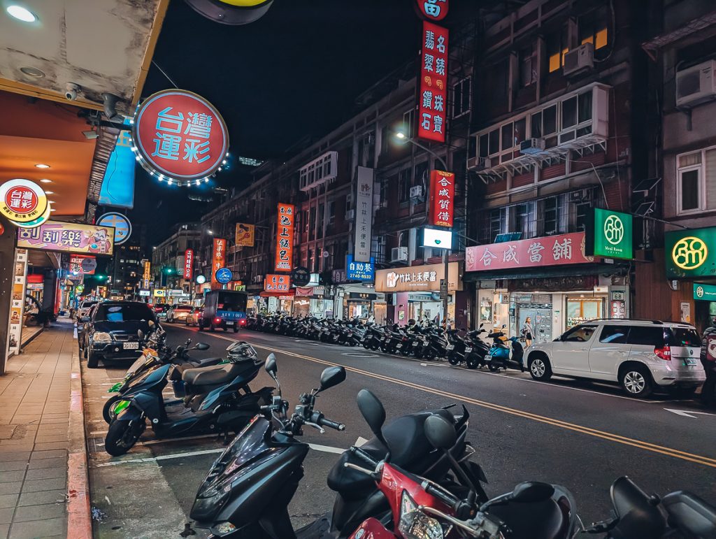 A Taipei street at night with lots of scooters parked on the side of the road under neon signs with Chinese writing.
