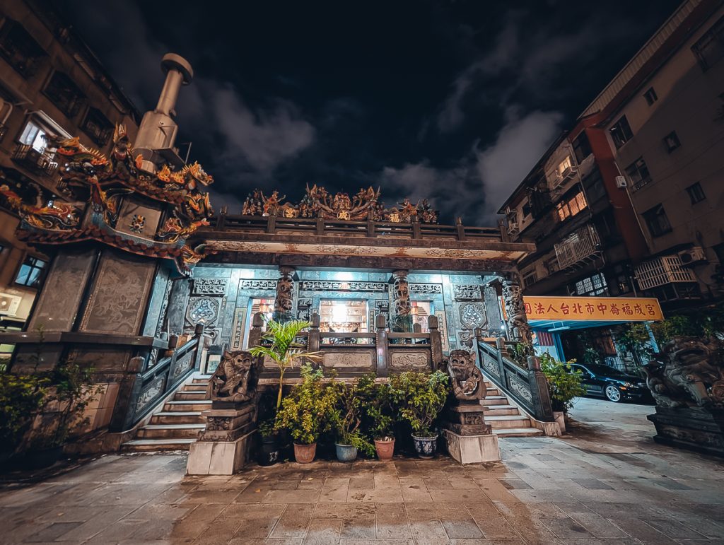 A small temple at night with potted plants and statues at the front.