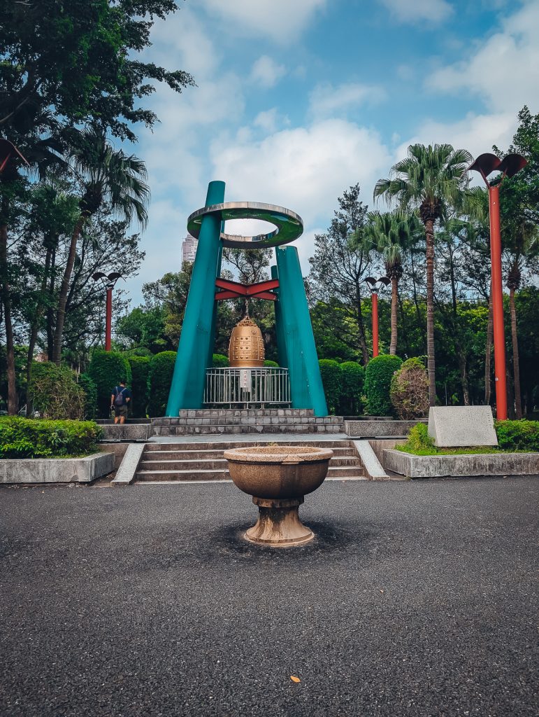 A blue and silver structure with a gold bell hanging from orange struts in a city park.
