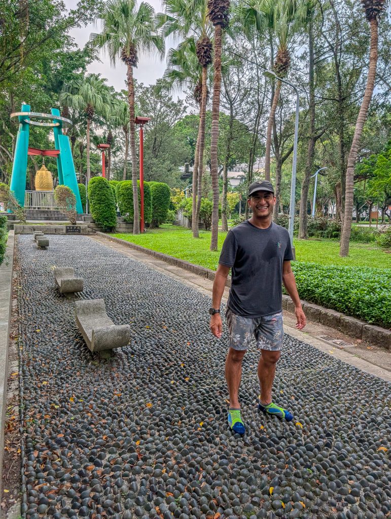Ganesh standing on a lumpy rock path in his socks grinning at the camera, with lush greenery and a blue and gold sculpture behind him.