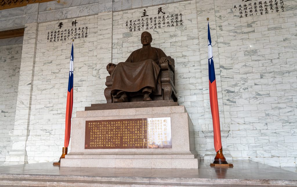 A bronze statue of a seated man on a marble plinth with Chinese writing on the walls and plinth beneath him.