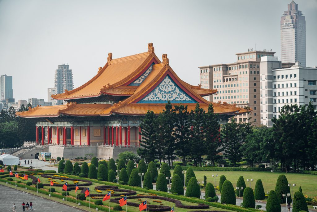 A grand pagoda-style building with an orange roof surrounded by manicured lawns and skyscrapers in the background.