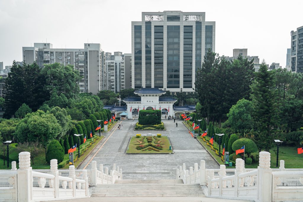 The view from on top of white stairs leading down to formal gardens and a temple-like gate, with skyscrapers in the background.