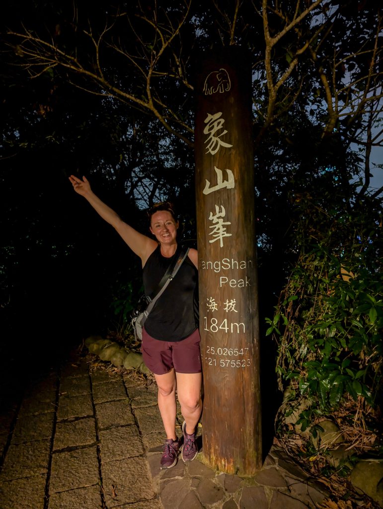 Sooz posing next to a wooden post with information about the Elephant Mountain peak on it, at night.