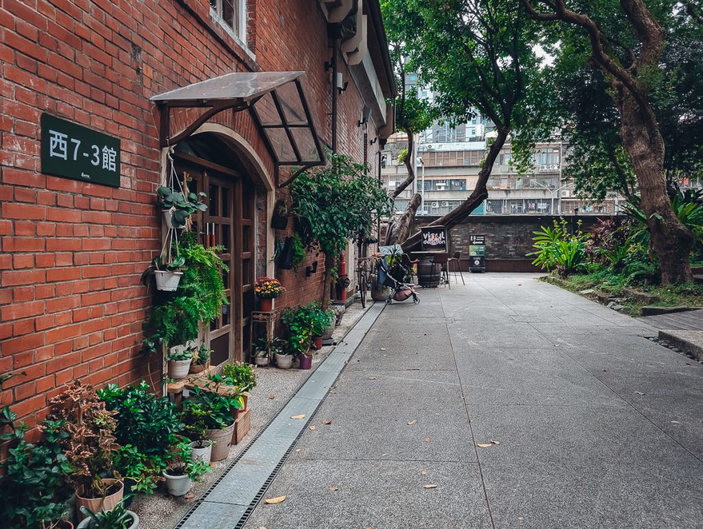 A brick building with lots of potted plants around the entrance and a concrete area in front.