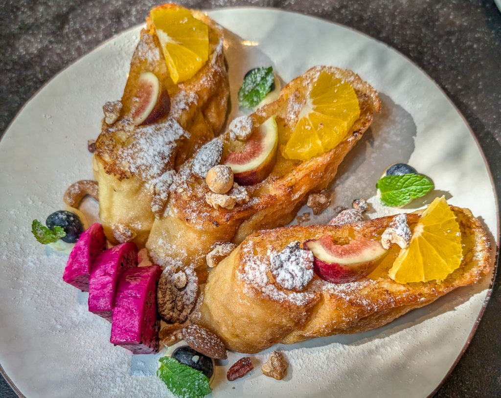 An overhead shot of a white plate of French toast with powdered sugar and colorful fruits.