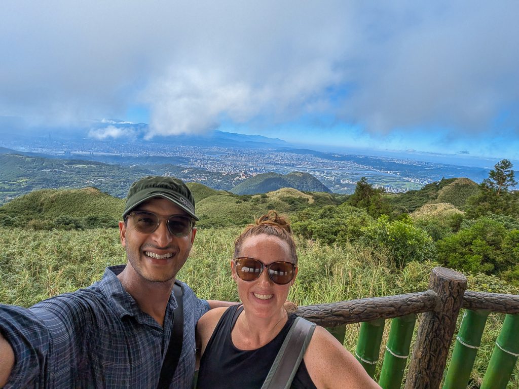 Sooz and Ganesh taking a selfie on top of a green mountain with a city and clouds behind them.