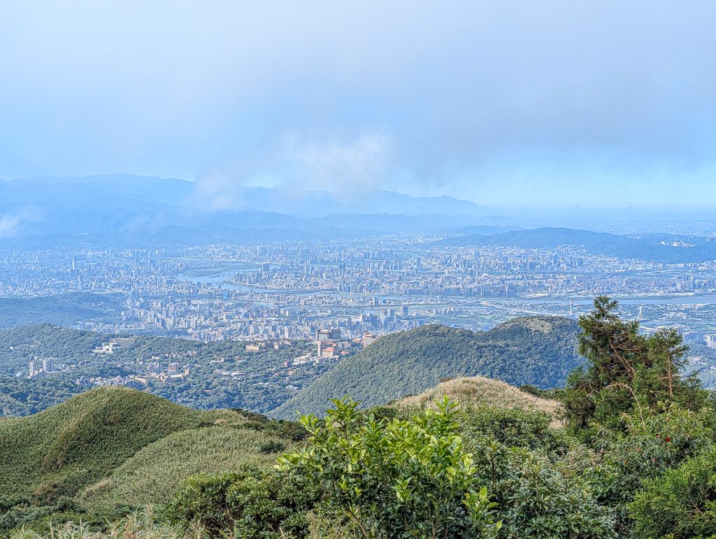 A view of a sprawling city as seen from a green hilly area with very misty clouds in the background.