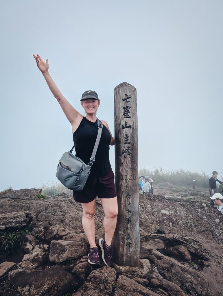 Sooz posing with a wooden mountain peak post on rocks with a misty sky behind.