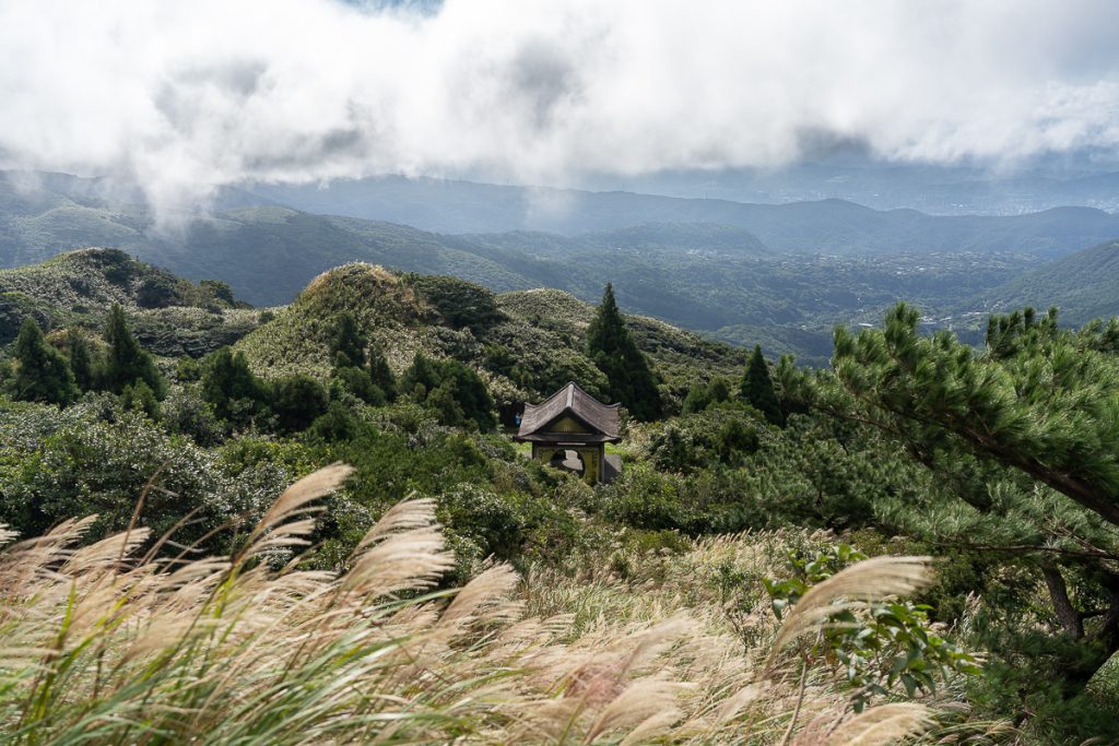 A small temple-like structure surrounded by greenery on a mountain, with more mountains and clouds in the background.